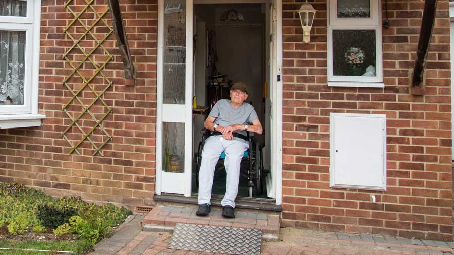 An elderly man sits in a wheelchair in the doorway of his home