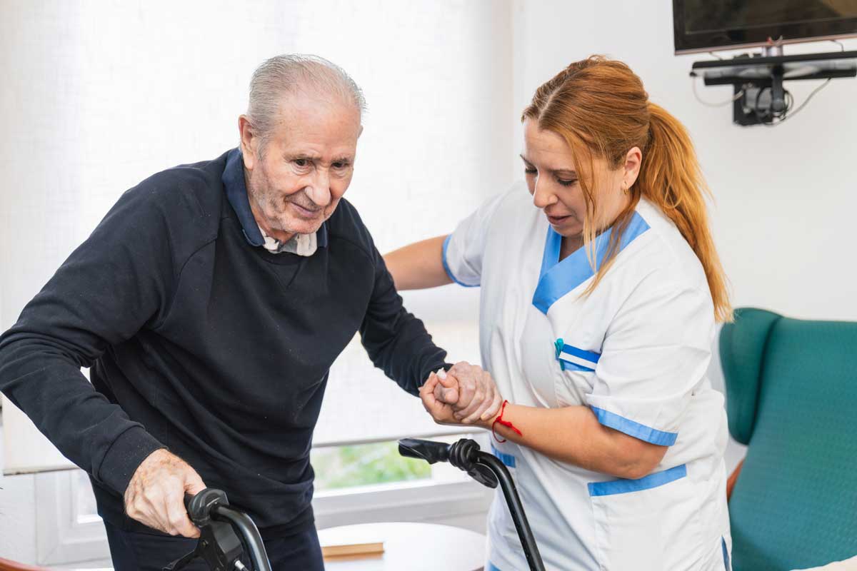 A care worker helps and older man to his walking frame