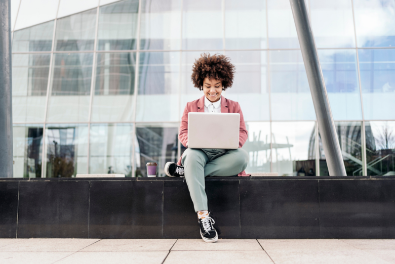 A young business woman sitting with a laptop on a step outside a corporate looking building, with a coffee cup next to her.