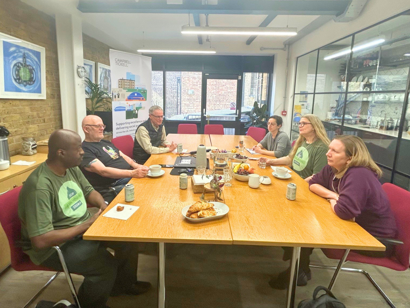 Group of people sitting around a table in a meeting room. There is a Campbell Tickell banner in the background. Two of the people at the table are wearing Stop the Stigma t-shirts.