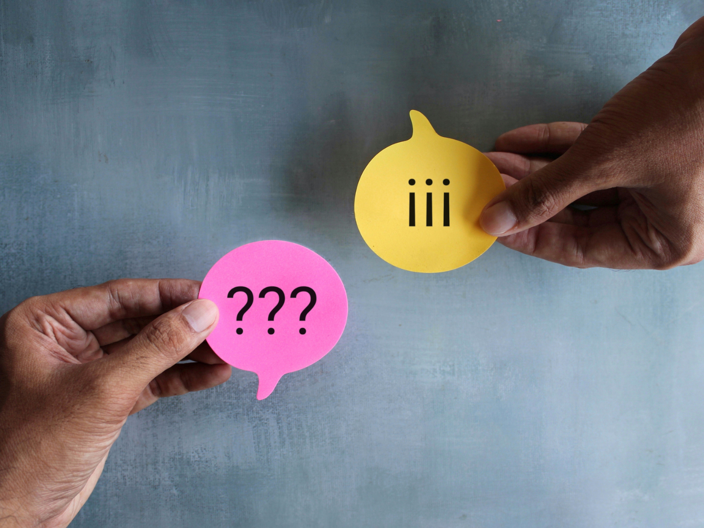 Two hands hold colourful speech-bubble cutouts against a grey background - one pink bubble with “???” and one yellow bubble with 